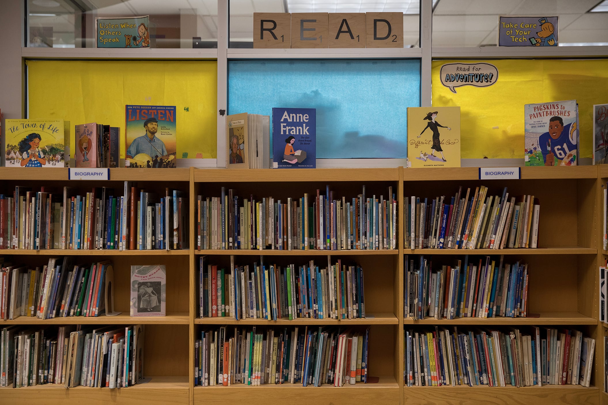 A bookshelf full of books line a wall with windows covered in yellow and blue paper above the bookshelf. There is a sign that says "READ" above the bookshelf.