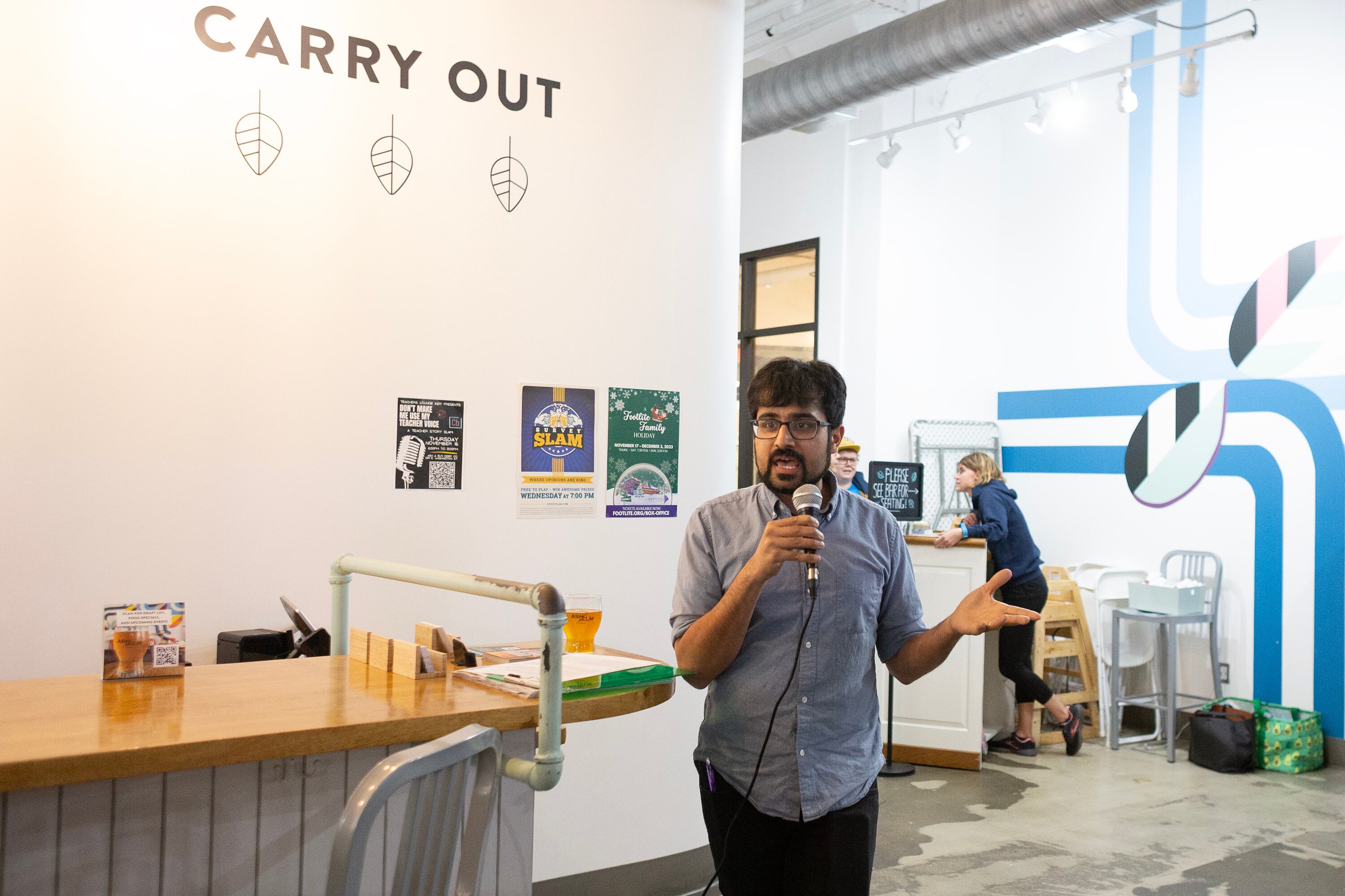 A man wearing a light grey shirt speaks into a microphone while a person stands in the background.