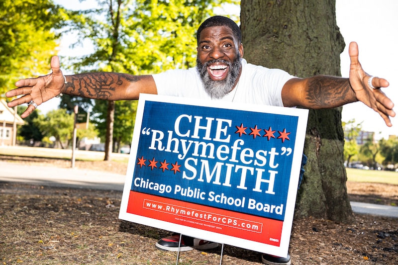 A man wearing a white t-shirt stands in front of an election sign outside.