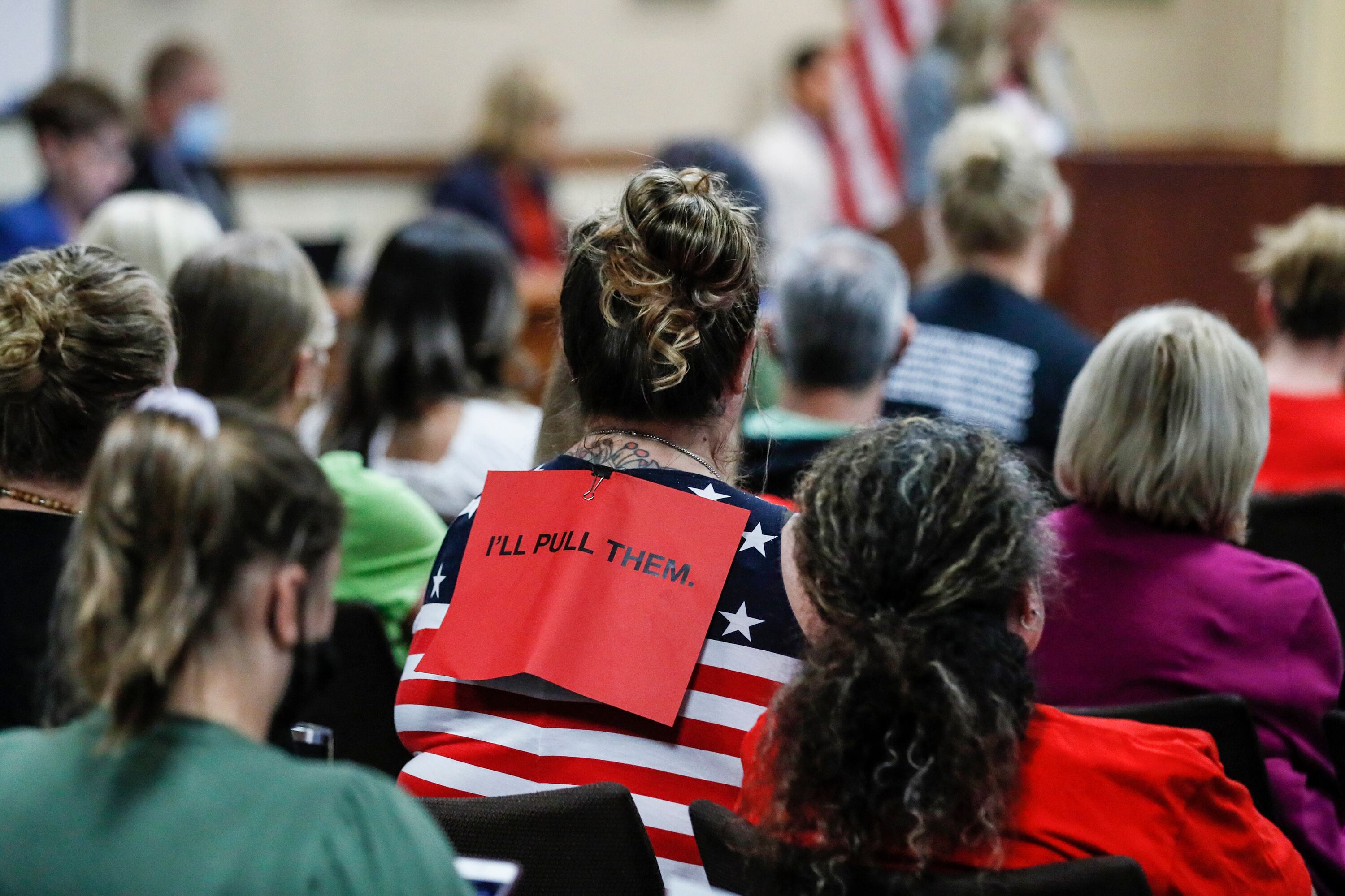 The back of people at a board meeting, with a red sign on the back of a person wearing an American flag shirt.