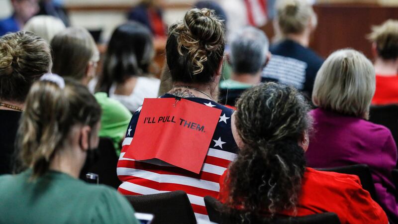 The back of people at a board meeting, with a red sign on the back of a person wearing an American flag shirt.