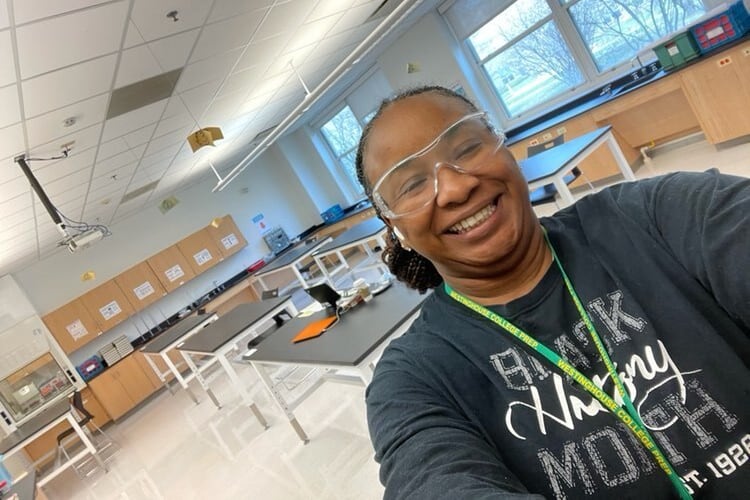 A woman wearing safety goggles, a school lanyard and a shirt that says “Black History Month” takes a selfie in her chemistry classroom.