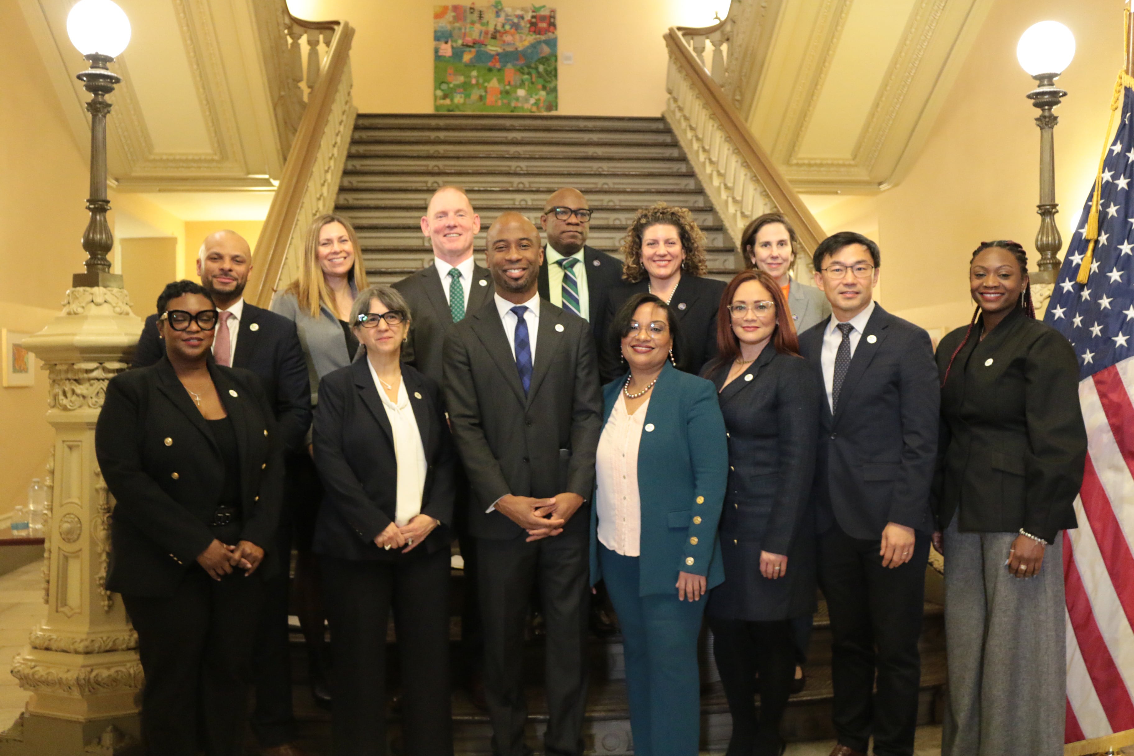A photograph of a group of people in suits standing at the base of a large set of stairs posing for a portrait with part of an American flag showing on the right side of the image.
