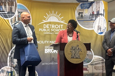 A white, bald man in a suit holds a blue gift bag while standing next to a Black woman in a peach suit speaking from behind a wooden podium and a microphone and both are in front of a yellow banner.