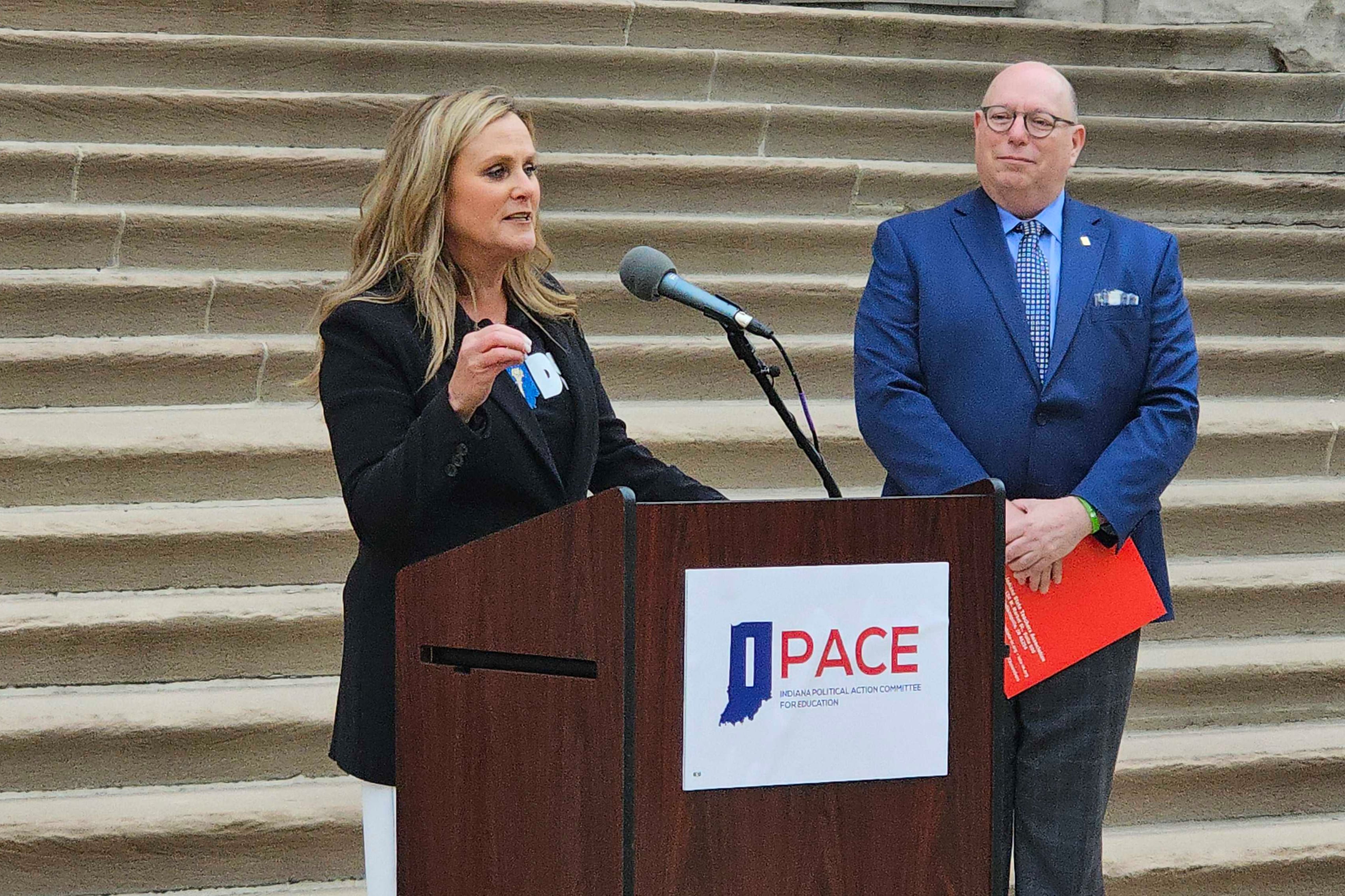 A woman with medium blond hair stands behind a wooden podium with a person wearing a blue suit and a set of stone stairs in the background.