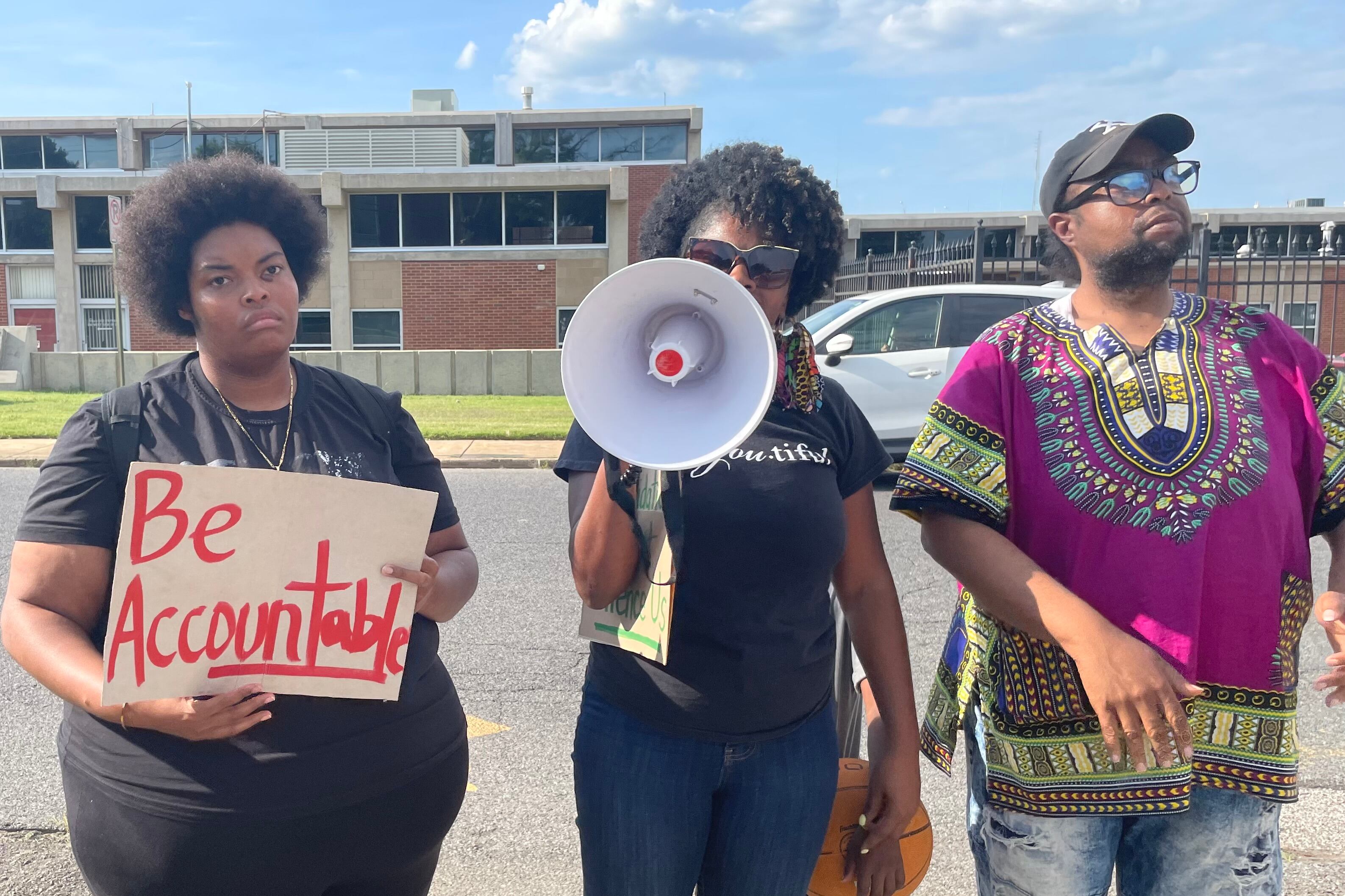 Three people standing outside a building. One has a sign that says “Be Accountable”; One has a megaphone. One is wearing a magenta shirt.