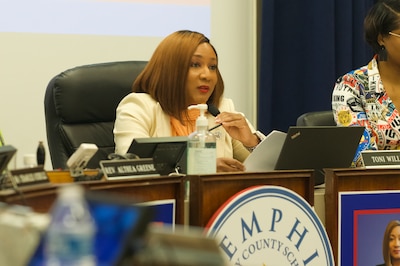 A woman with auburn hair and a white blazer sits in front of a laptop and adjusts her microphone to speak.