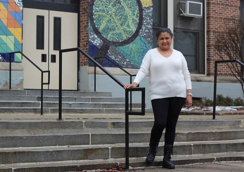 A woman wearing a white sweater stands outside the front of a school building on the steps.