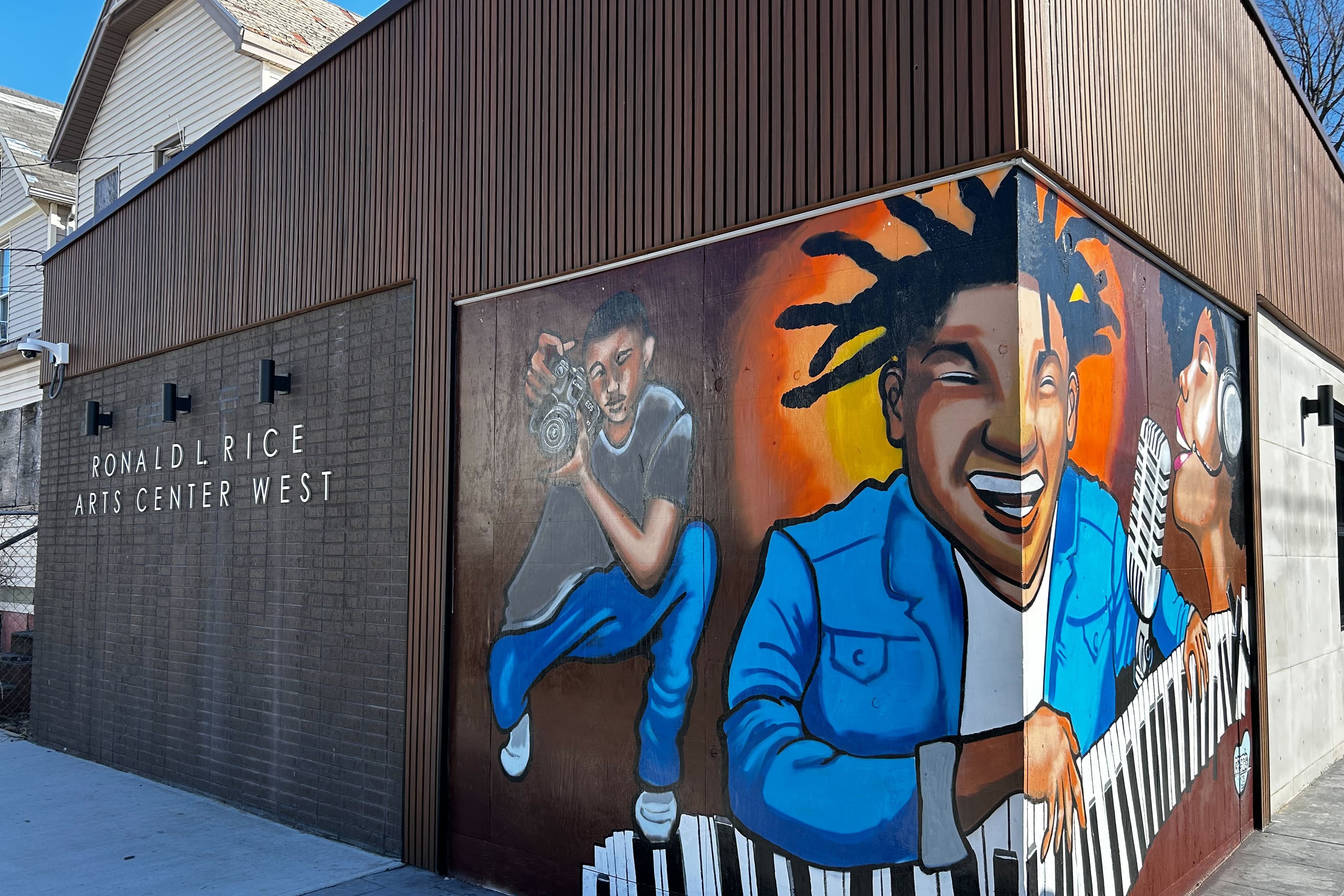A large mural showing three people playing the piano, singing and using a camera on the corner of a building.