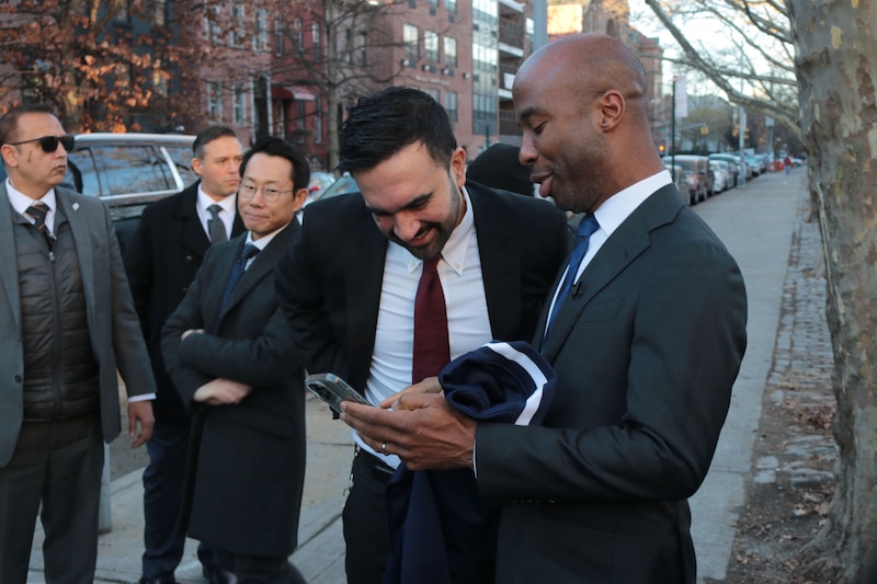 A photograph of two men in suits talking with each other outside next to a group of adults.