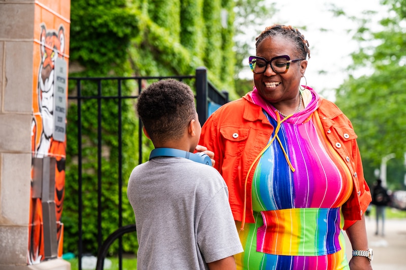 A Black woman in a colorful dress speaks to a young student outside of a school building.