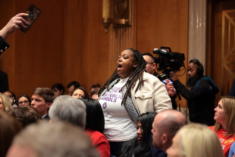 A woman with long dark hair stands and speaks amongst a group of people sitting in rows of chairs.