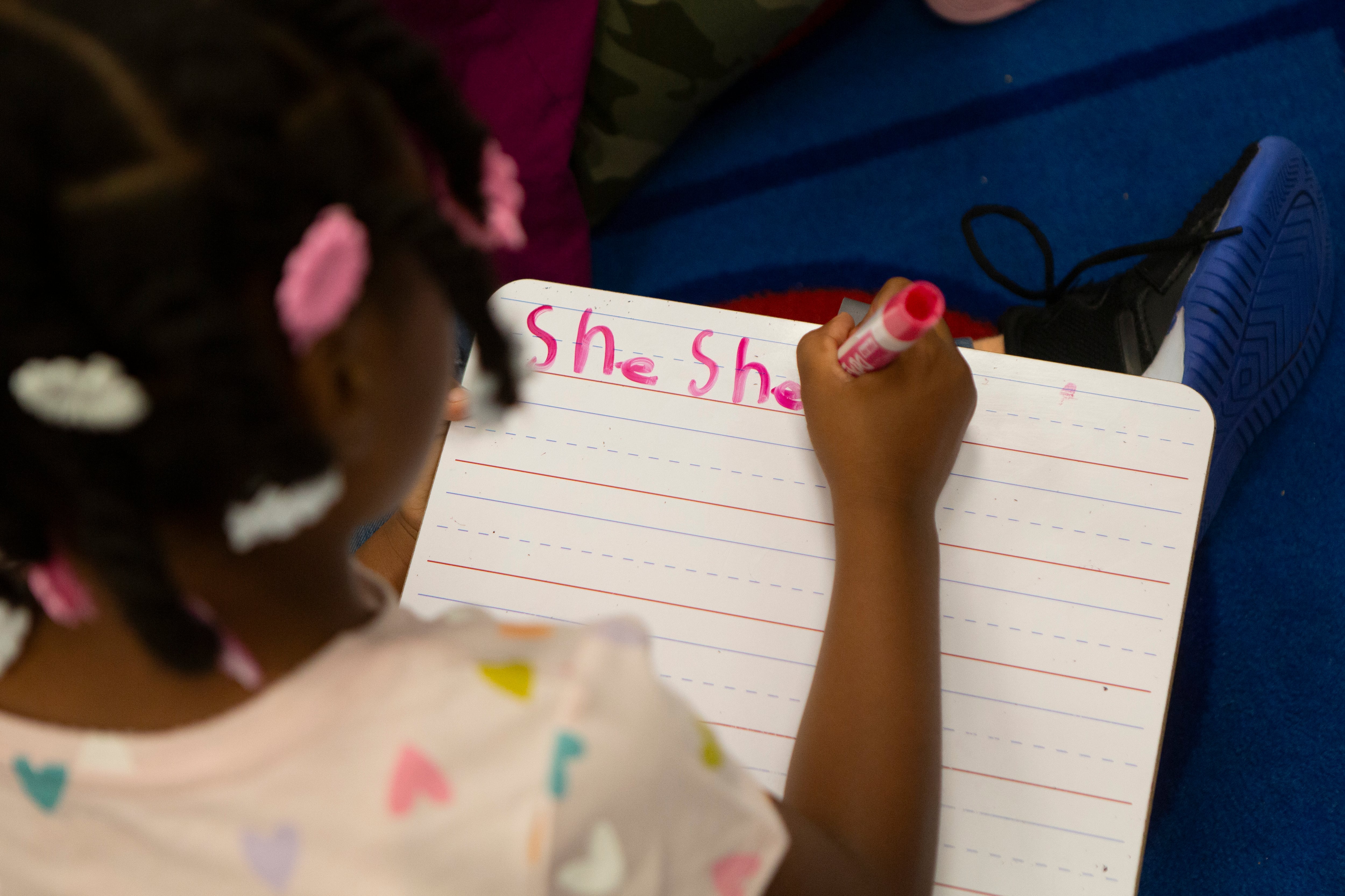 A view from above of a young child with braids and colorful beads in her hair practicing writing the word "she" on a dry erase board in red marker.