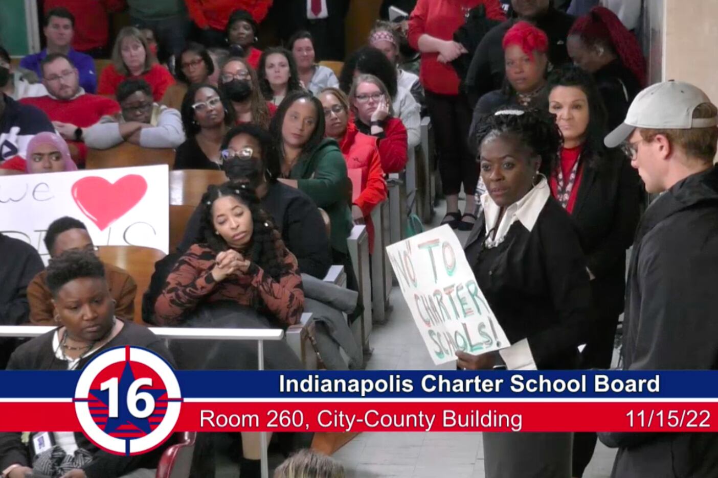 people in chairs at a meeting and one woman holds a sign that says no to charter schools.