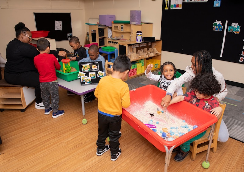 Two adults each sit at a small table with young students in a preschool classroom.