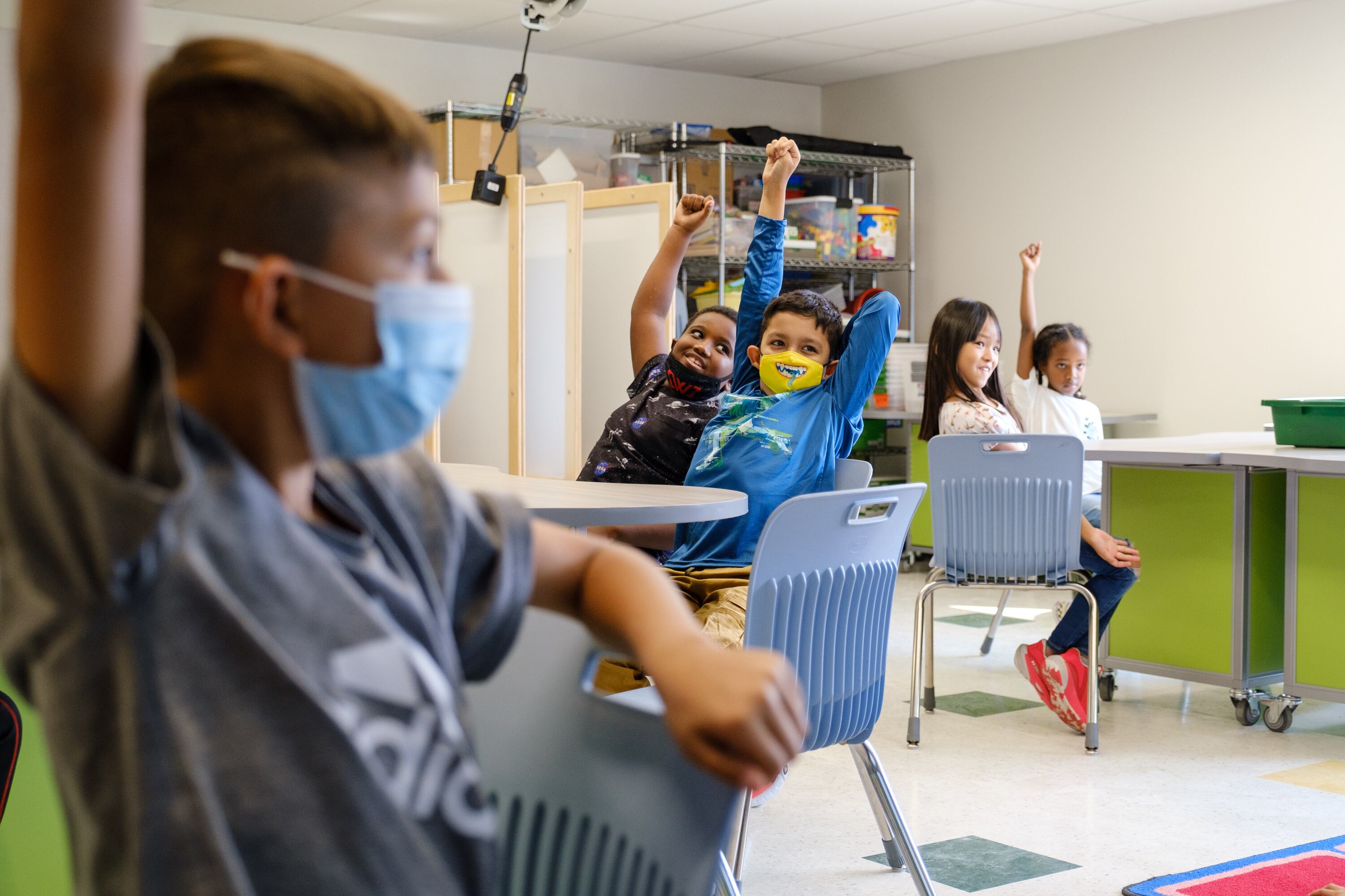 Elementary students, some wearing masks, sit in plastic chairs while raising their fists in the air during an activity in class at Virginia Court Elementary School in Aurora.