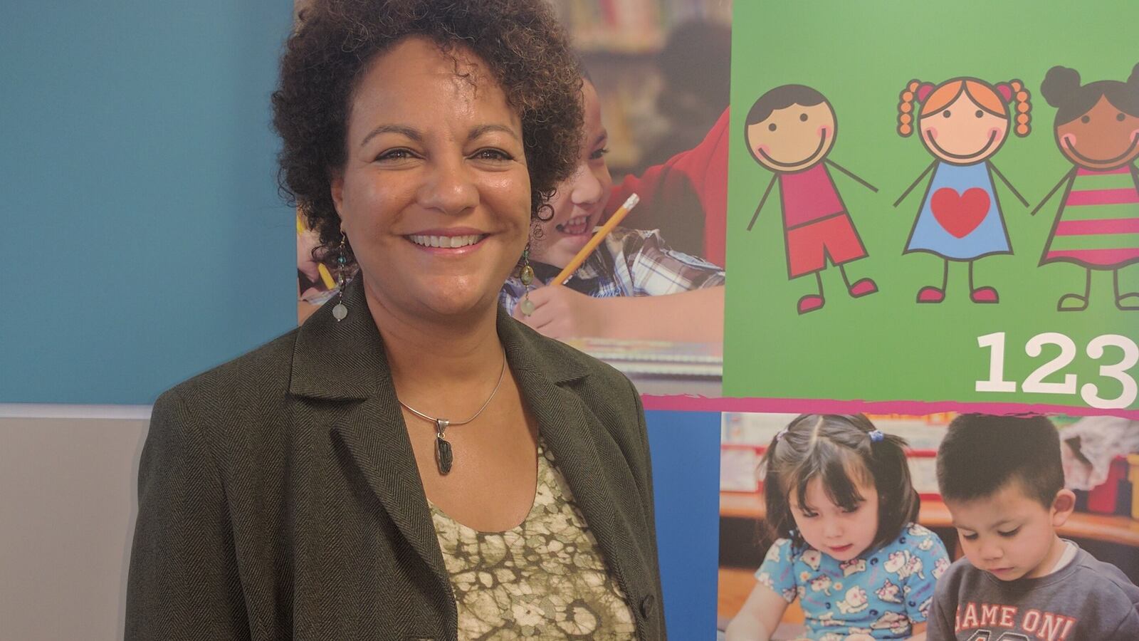A woman with short brown hair who is wearing a dark blazer and a necklace stands in front of a bulletin board with drawings and photos of children on it.