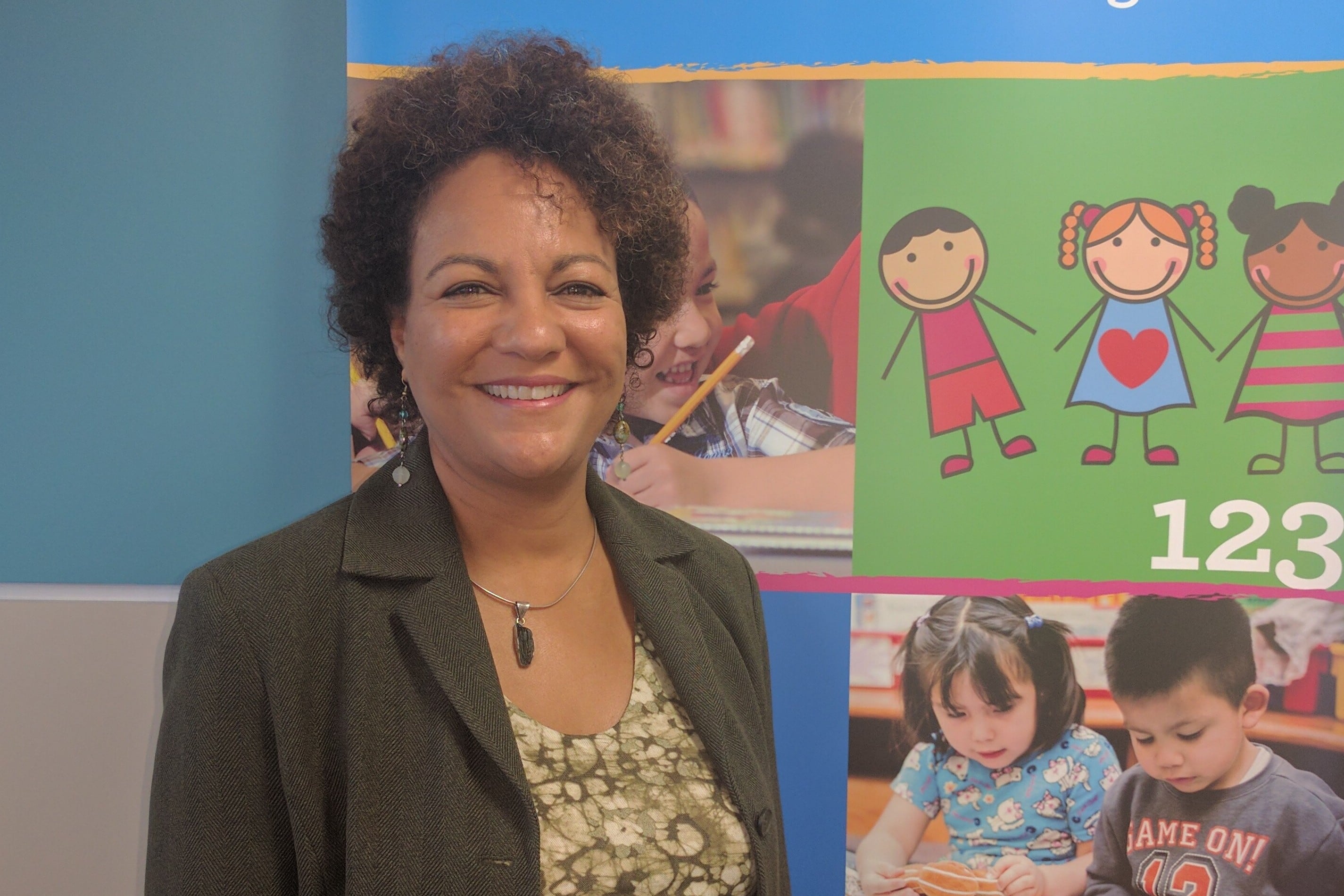A woman with short brown hair who is wearing a dark blazer and a necklace stands in front of a bulletin board with drawings and photos of children on it.