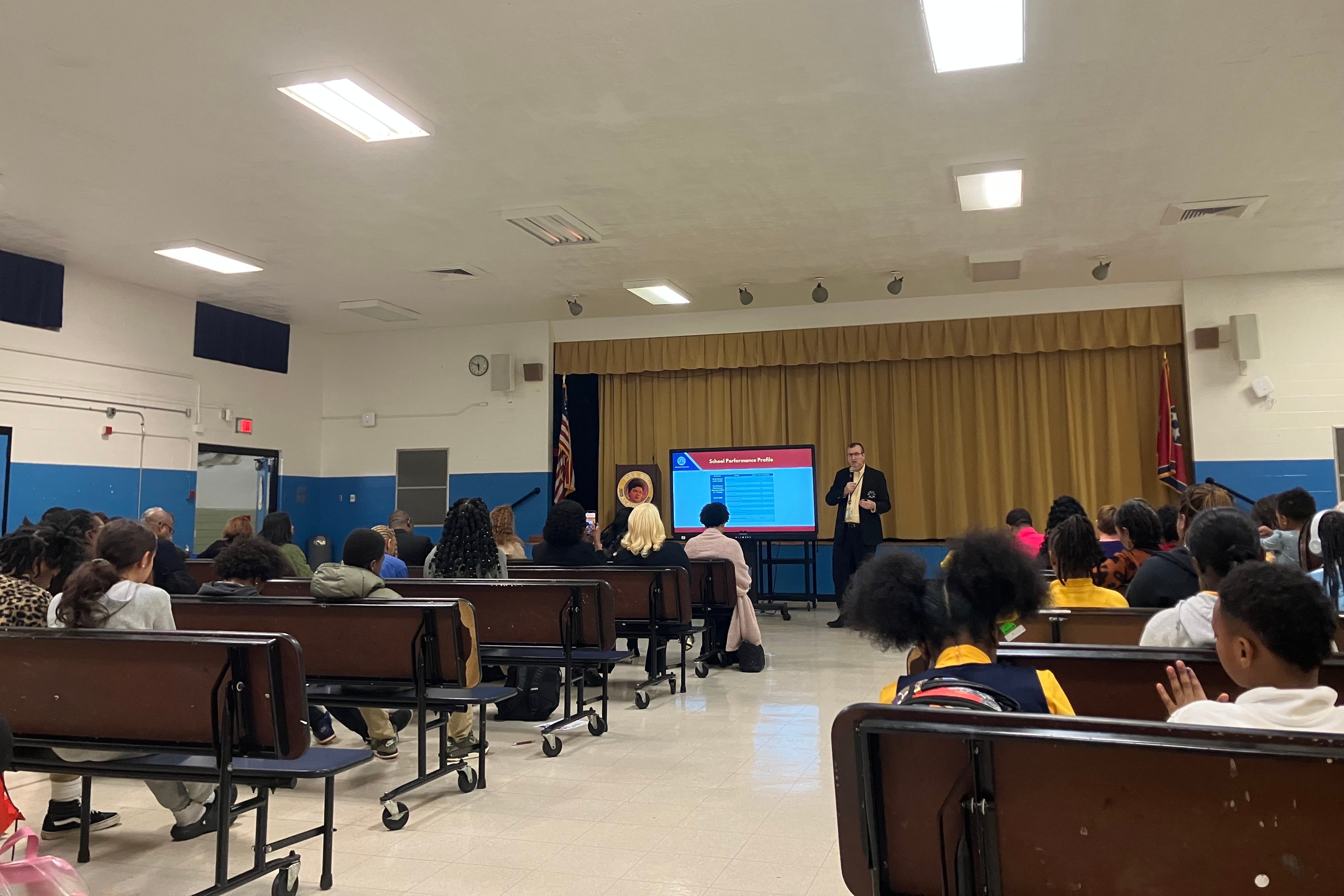 In a school auditorium, a man in a suit stands beside a lit-up TV screen in front of mustard yellow curtains. Parents sit in rows of benches facing him.
