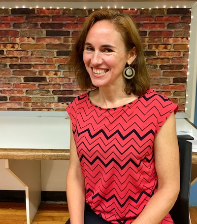A white woman with short light brown hair and wearing a red and black blouse sits for a portrait with a brick wall in the background.