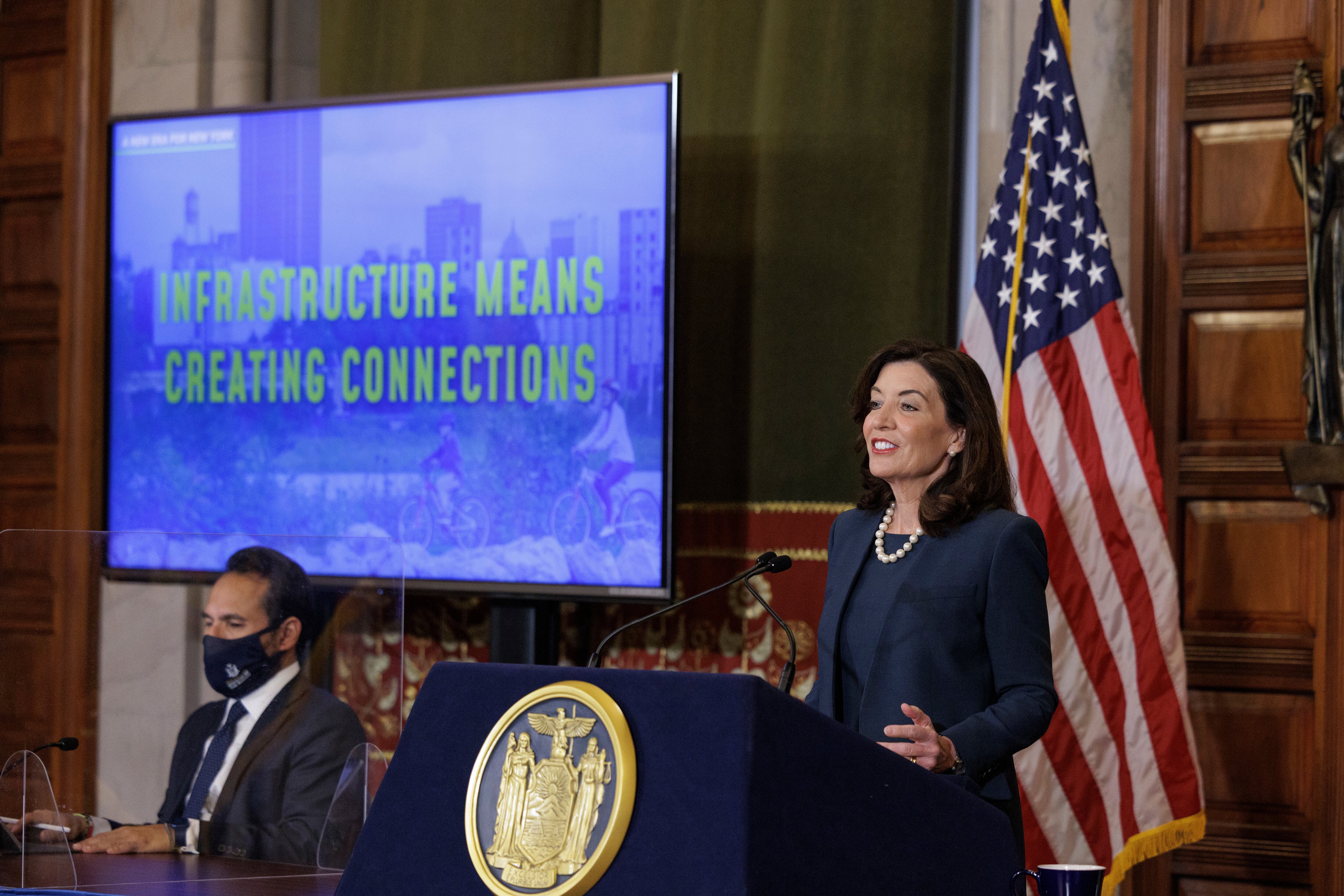 New York State Governor Kathy Hochul speaks at a podium in the State Capitol. There is a large TV playing a slideshow for the state’s yearly budget presentation in the background.
