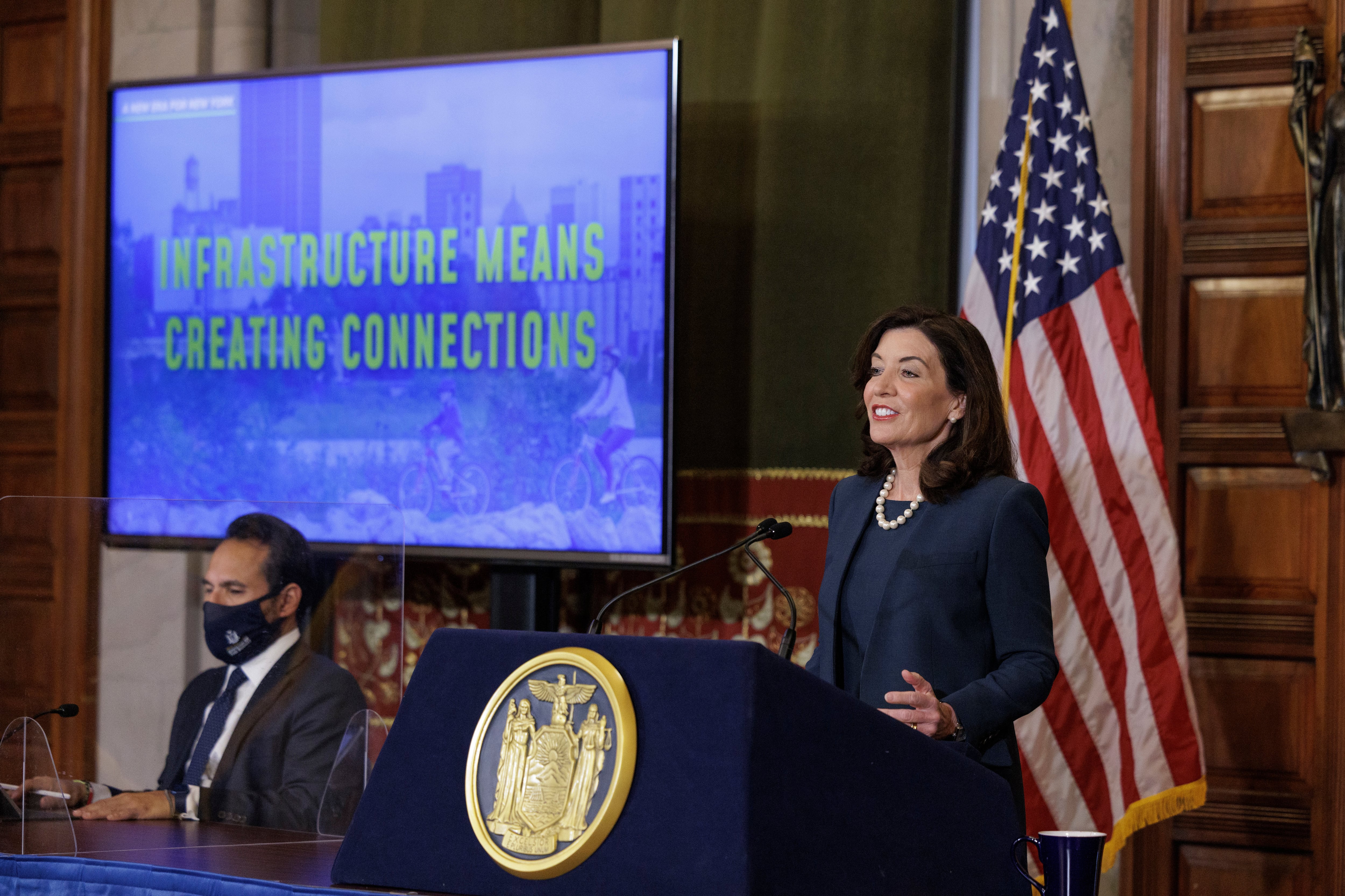New York State Governor Kathy Hochul speaks at a podium in the State Capitol. There is a large TV playing a slideshow for the state’s yearly budget presentation in the background.