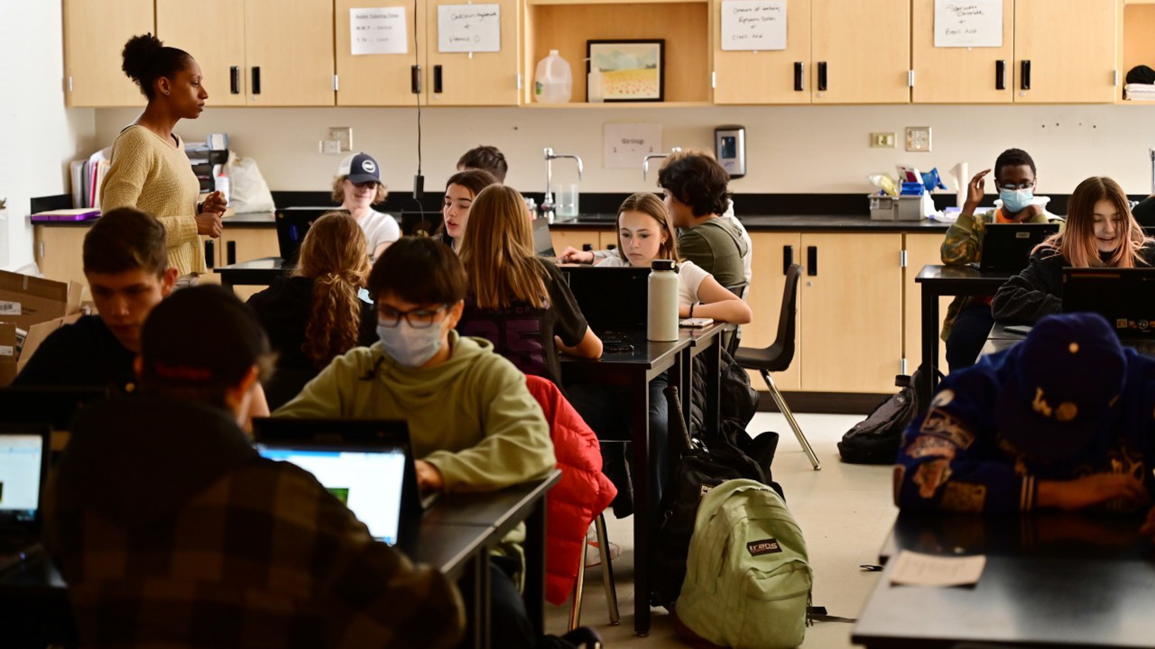 A high school teacher works with her students who are seated at tables in a science lab.