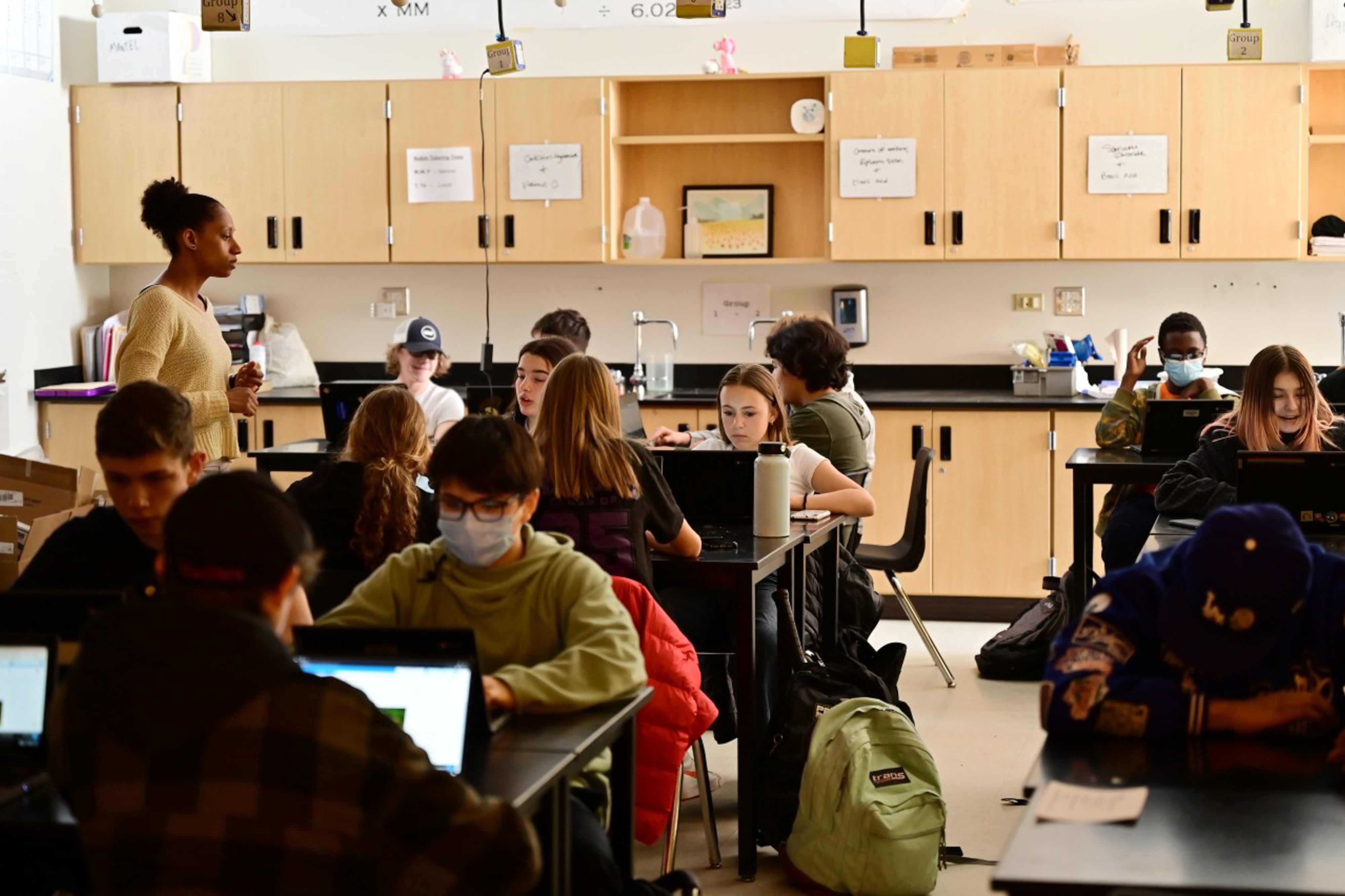 A high school teacher works with her students who are seated at tables in a science lab.