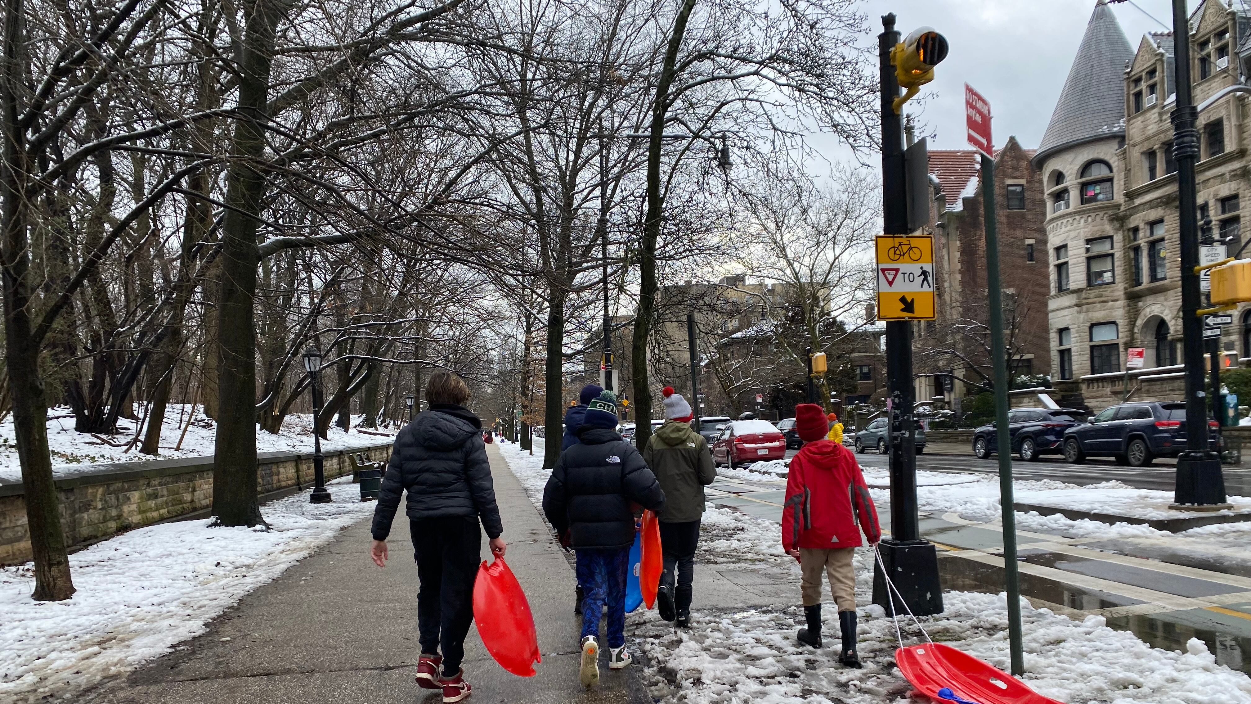 A group of children in winter gear with sleds walking on a sidewalk without much snow.