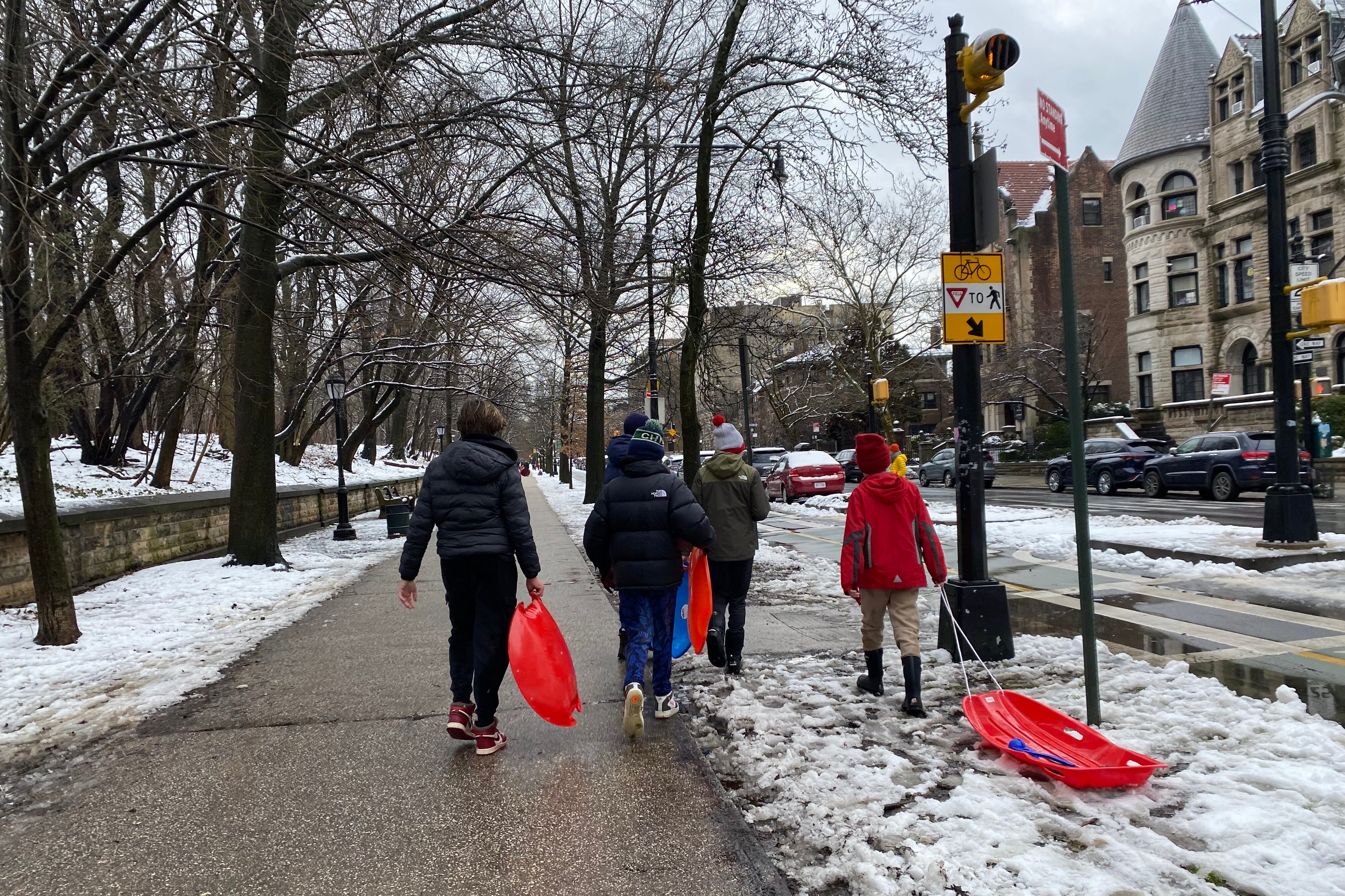 A group of children in winter gear with sleds walking on a sidewalk without much snow.
