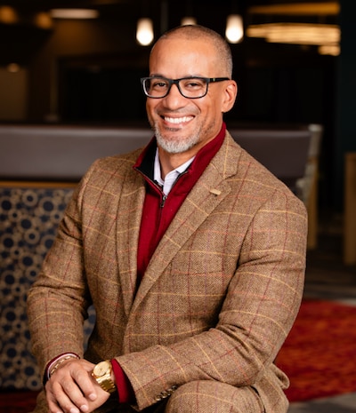 A photograph of a Black man wearing glasses and a brown suit with a smile posing for a portrait.