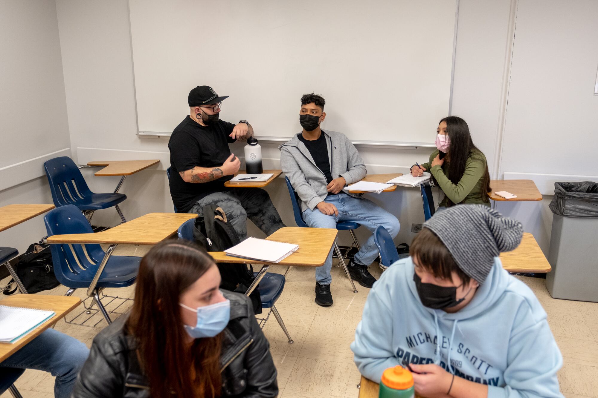 Five college students sit at desks in a classroom with white walls and a white marker board in the background
