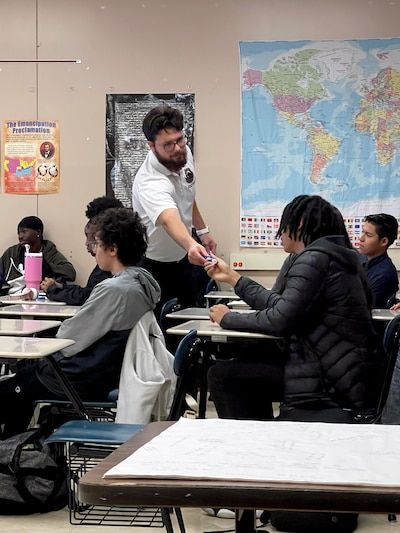 A teacher stands next to a group of students sitting at their desks in a classroom.