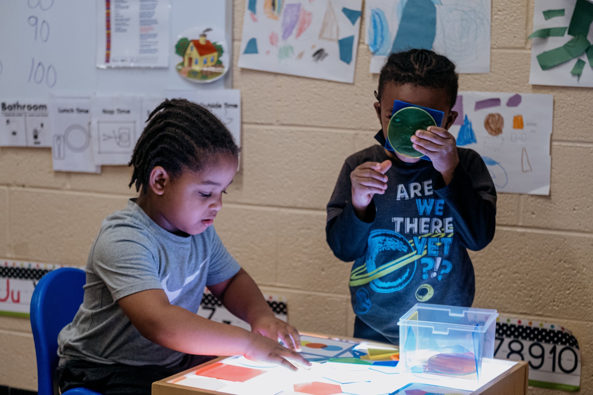 One young child sits at a table, while another stands nearby with classroom artwork hanging on the wall behind them.