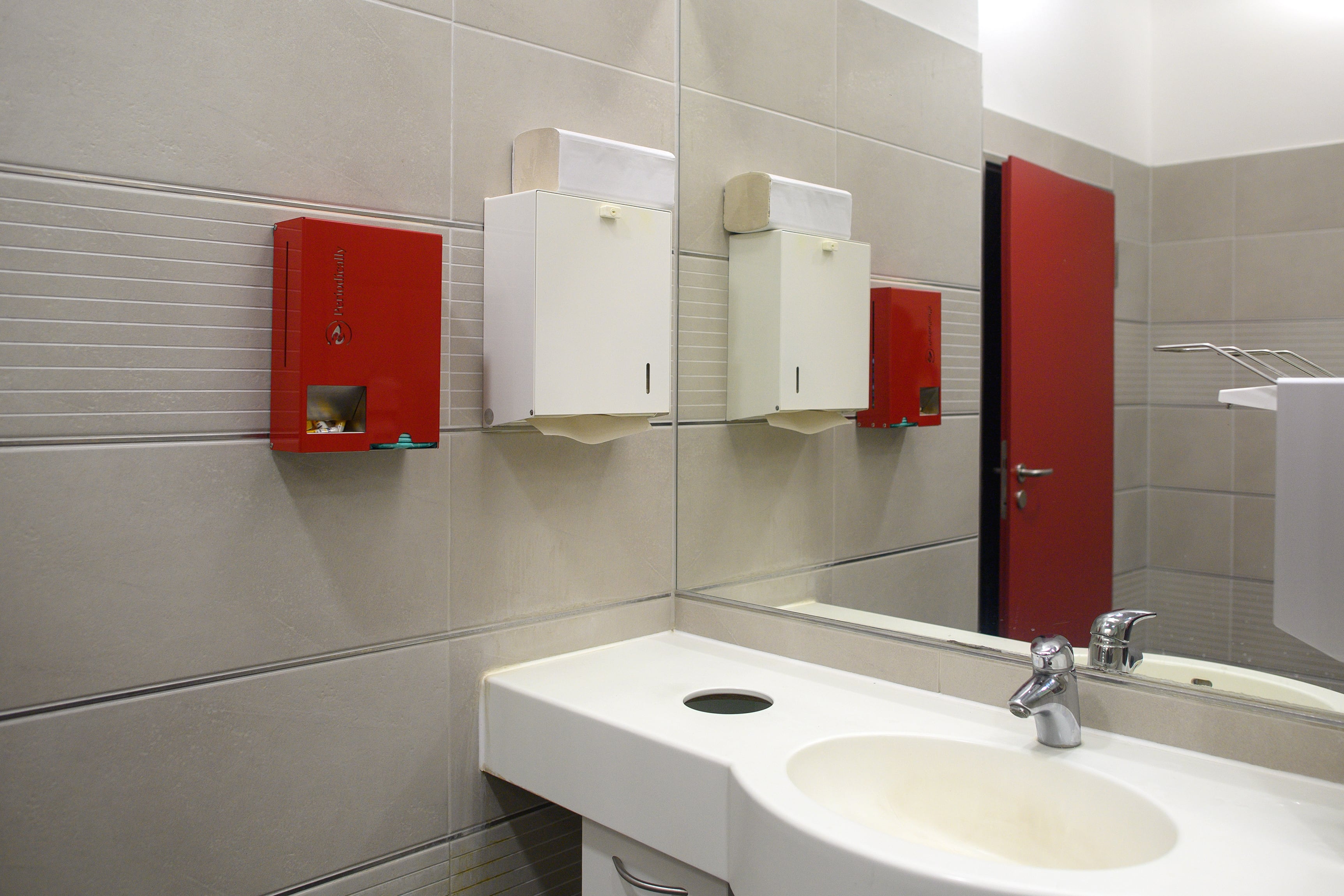 Red and white tampon and sanitary napkin dispensers on the side of a bathroom wall with a sink and a mirror.