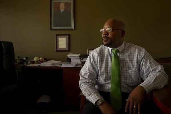 Aurora Superintendent Rico Munn, wearing a green tie and glasses, poses for a portrait while seated. A portrait of Supreme Court Justice Thurgood Marshall hangs on the wall.