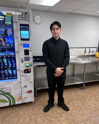A high school student in an all black suit poses for a portrait in a cafeteria.