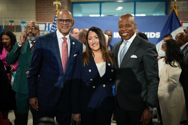 Three adults in suits stand side by side posing for a photo with a group of people behind them.
