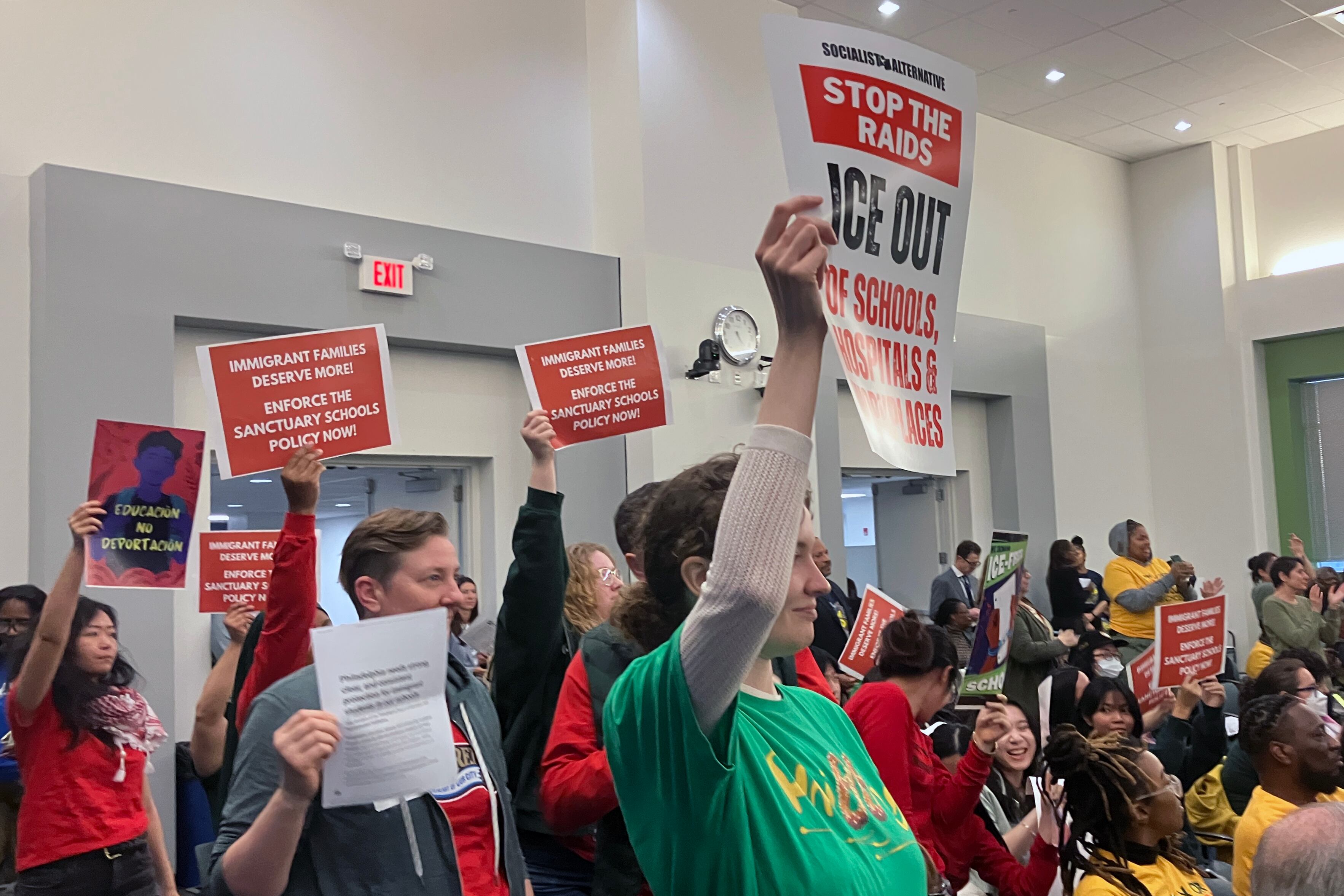 A group of community members hold up protest signs in a conference room.