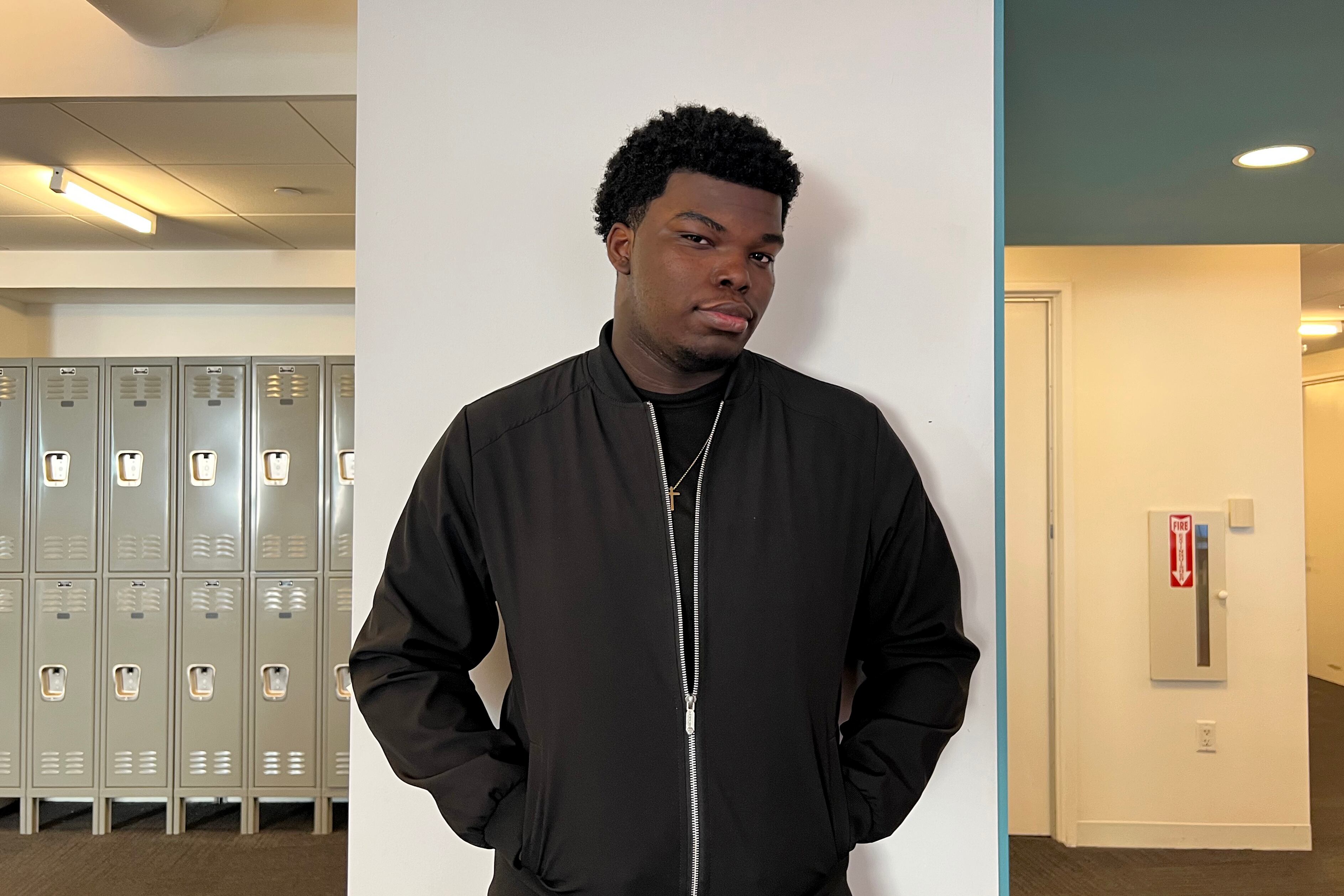 A photograph of a young Black man wearing a black zip up sweater posing inside of a school hallway.