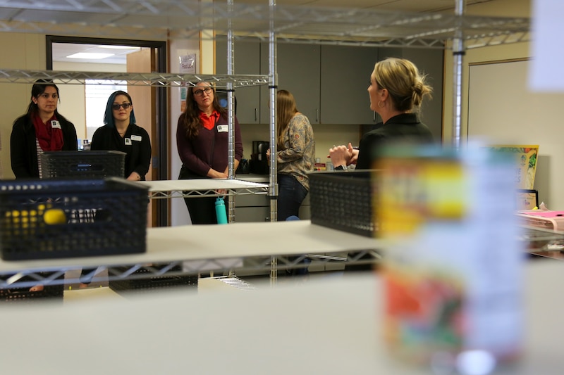 A group of people are seen through a gap in a metal shelf in a room with a can of food.