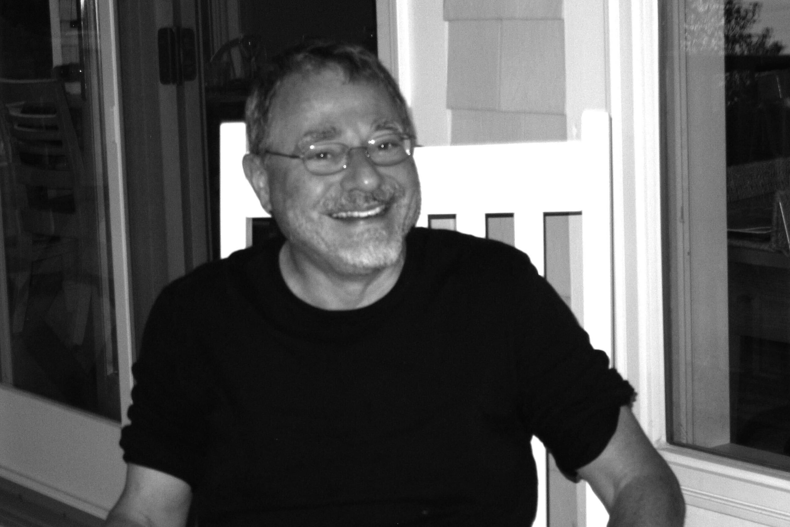 A man with glasses and a black t-shirt smiles at the camera from his front porch.