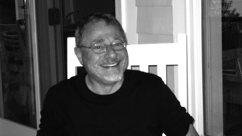 A man with glasses and a black t-shirt smiles at the camera from his front porch.