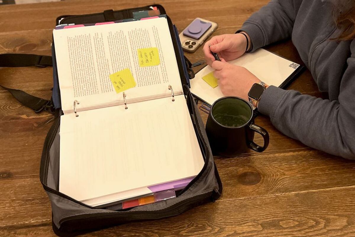A person is sitting at a table holding a pen, but only their hands are in view. They have a binder full of papers, a coffee cup, and a phone in front of them.