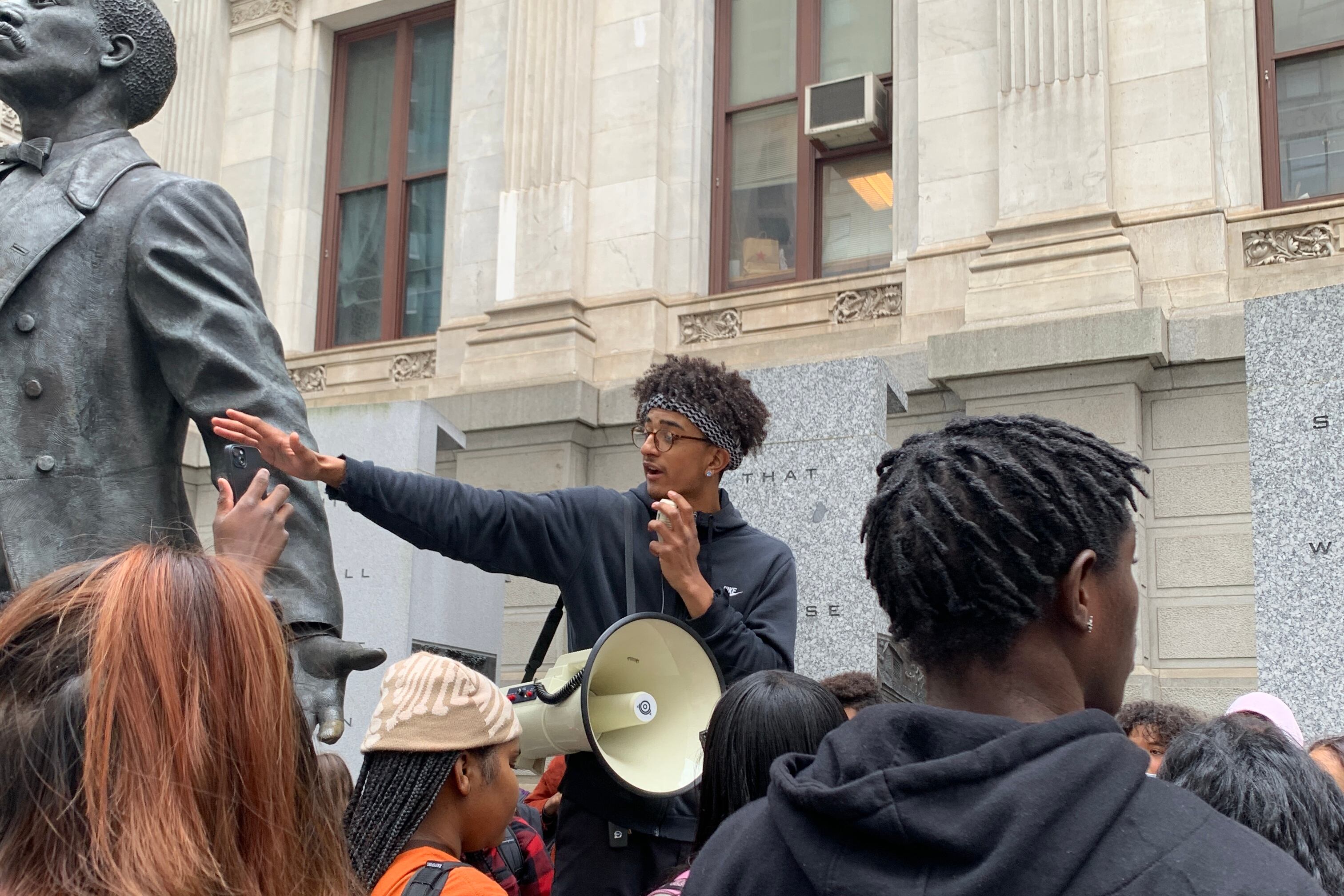 A person wearing a bandana, a dark shirt, and dark glasses gestures with his right hand as he and others stand in front of a stone building. A statute is to the person’s right.