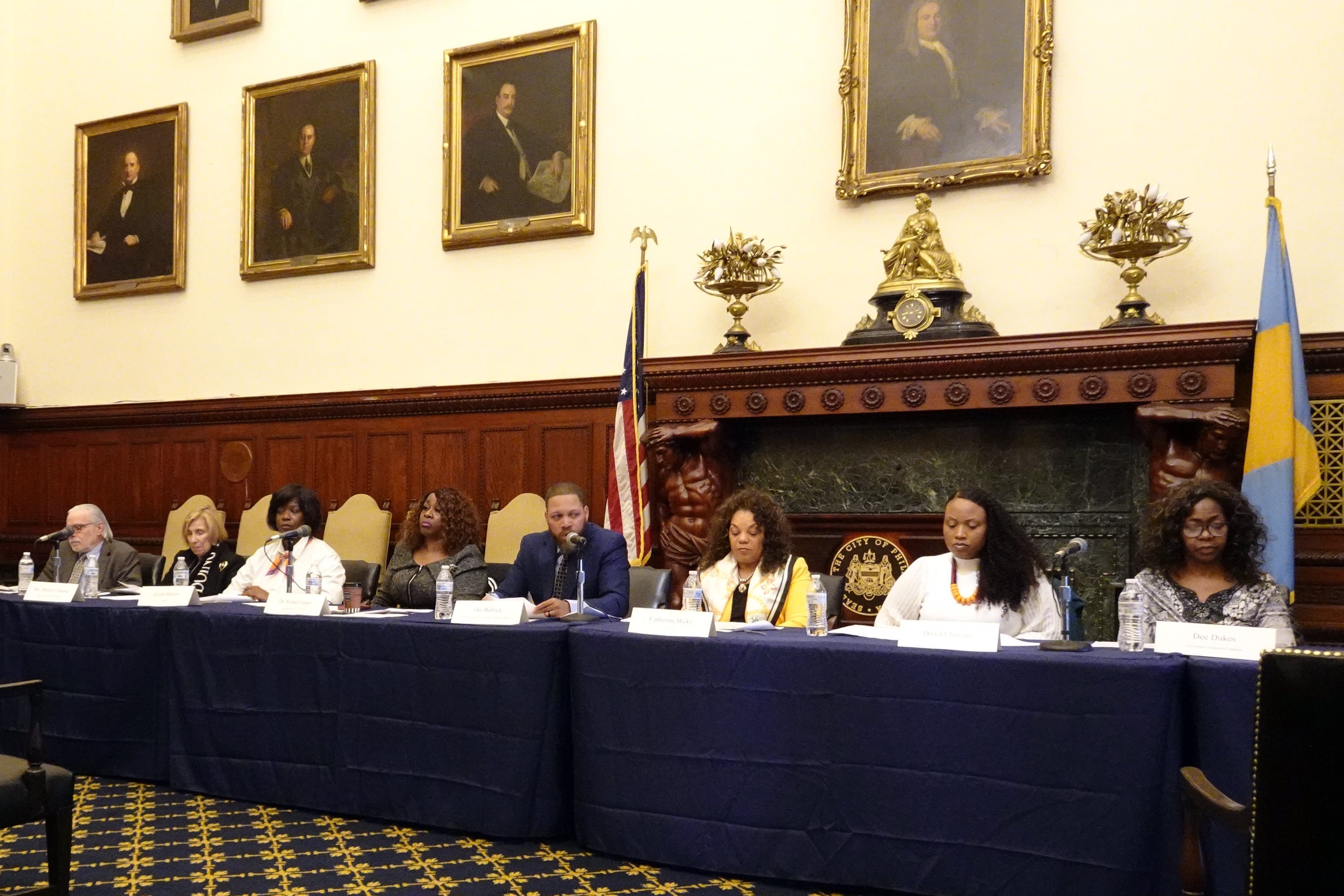 A group of people sit in a row at a long table with portraits on the wall and a flag in the background.