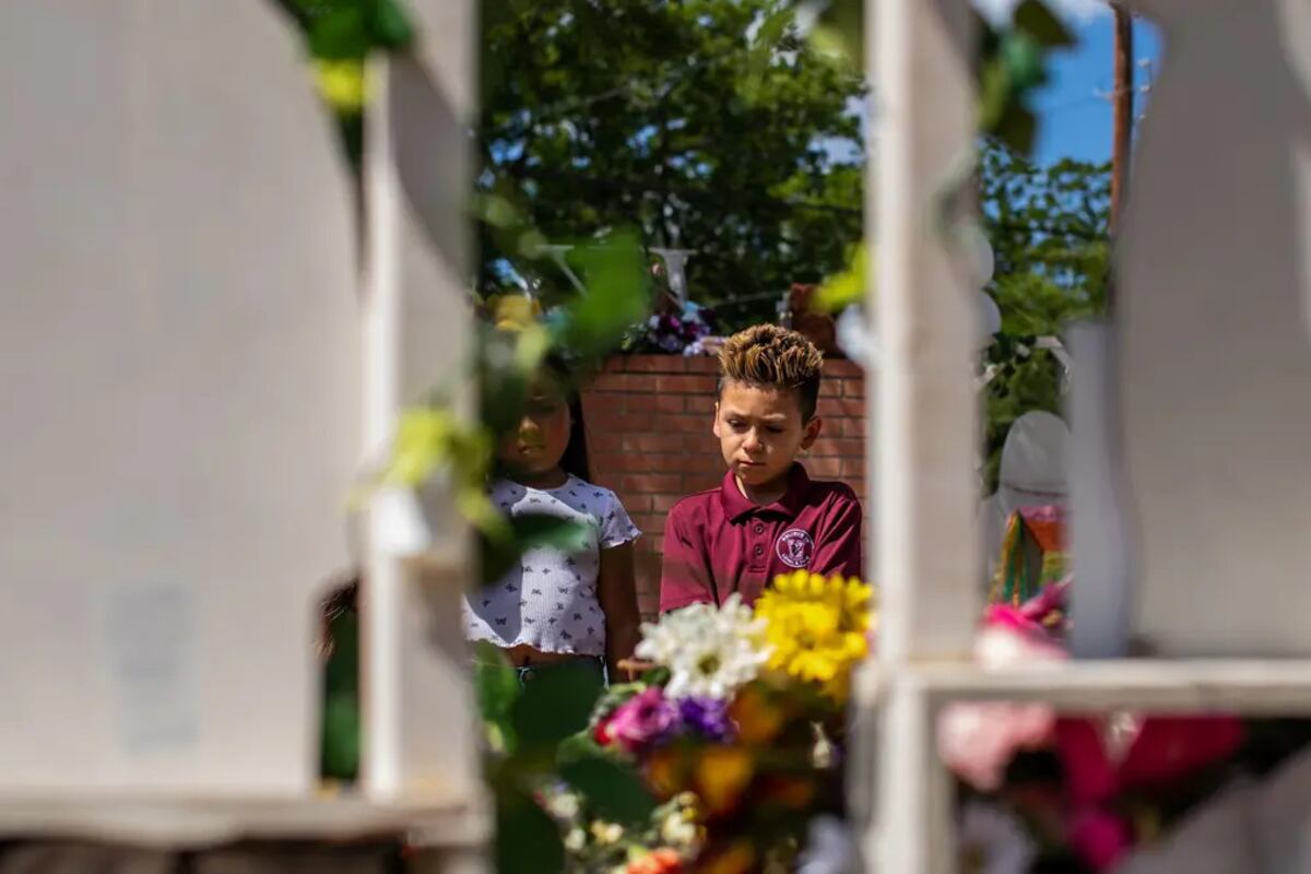 A young boy with spiky hair standing next to a young girl as they look down at flowers and balloons, seen through a white fence.