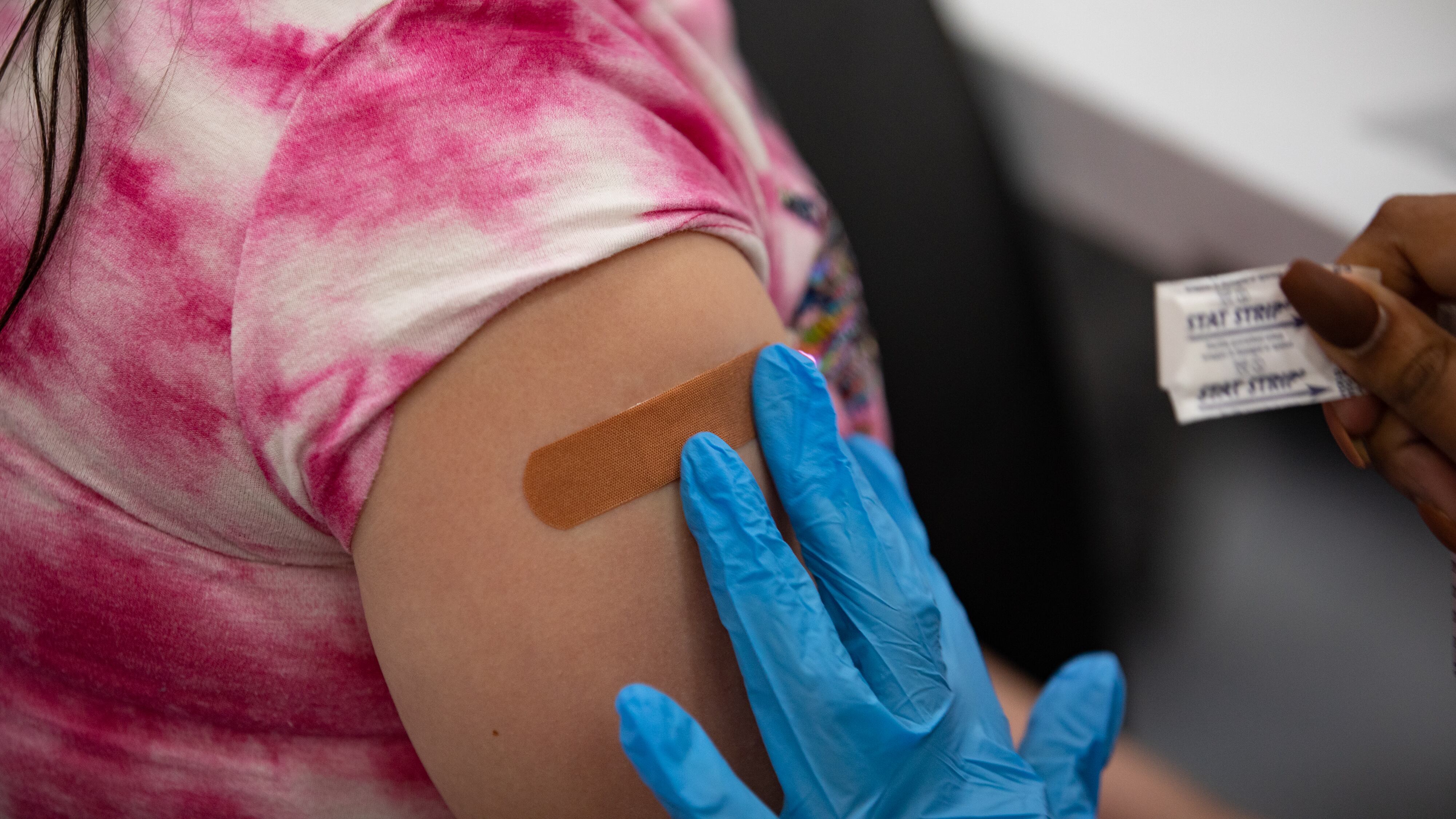 A gloved hand applies a flesh-colored bandaid to a child’s arm. The child is wearing a pink and white tie-dyed shirt rolled up to the shoulder. A few strands of dark hair are visible on the child’s shoulder.