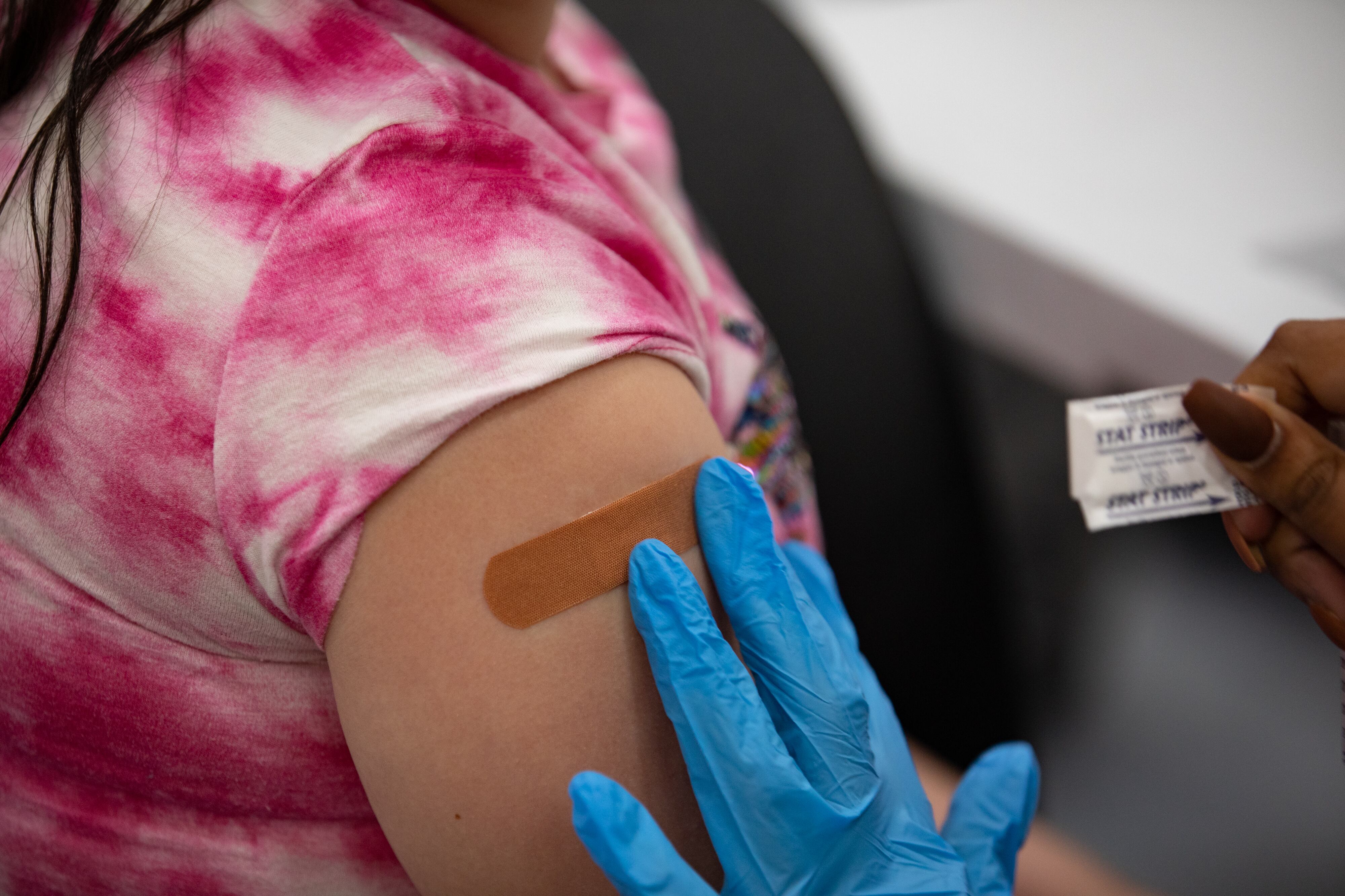 A gloved hand applies a flesh-colored bandaid to a child’s arm. The child is wearing a pink and white tie-dyed shirt rolled up to the shoulder. A few strands of dark hair are visible on the child’s shoulder.