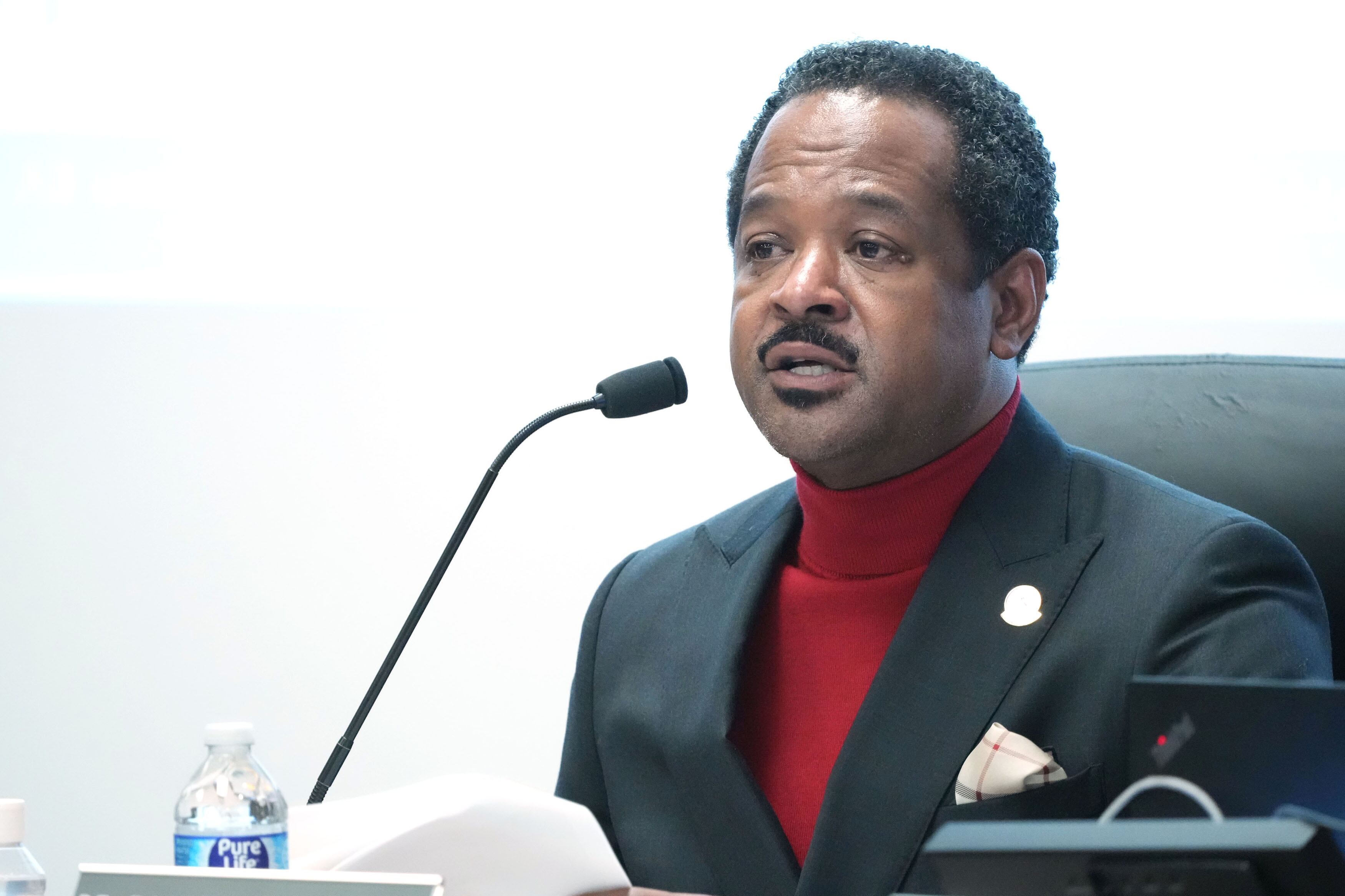 A Black man wearing a red shirt and a dark suit jacket speaks into a thin microphone while holding paper during a meeting.